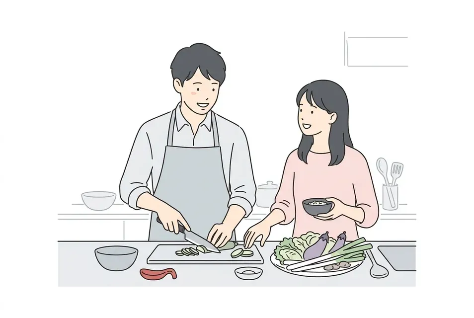 料理教室のキッチンカウンターで、エプロン姿の男女が笑顔で話しながら一緒に下ごしらえをする様子。